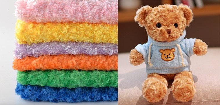 Stacked rolls of colorful curly plush fabric beside a teddy bear wearing a blue hoodie with a bear face.