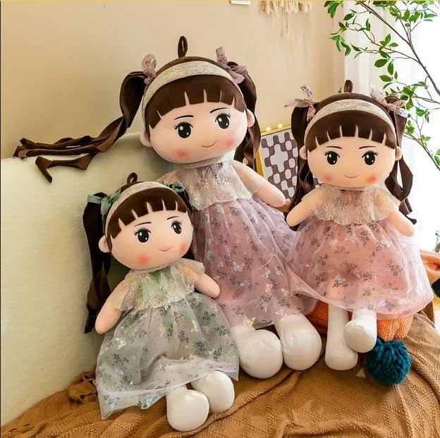 Three plush girl dolls with brown hair, wearing pastel-colored floral dresses and headbands, sitting on a couch with cozy decor and a potted plant nearby.