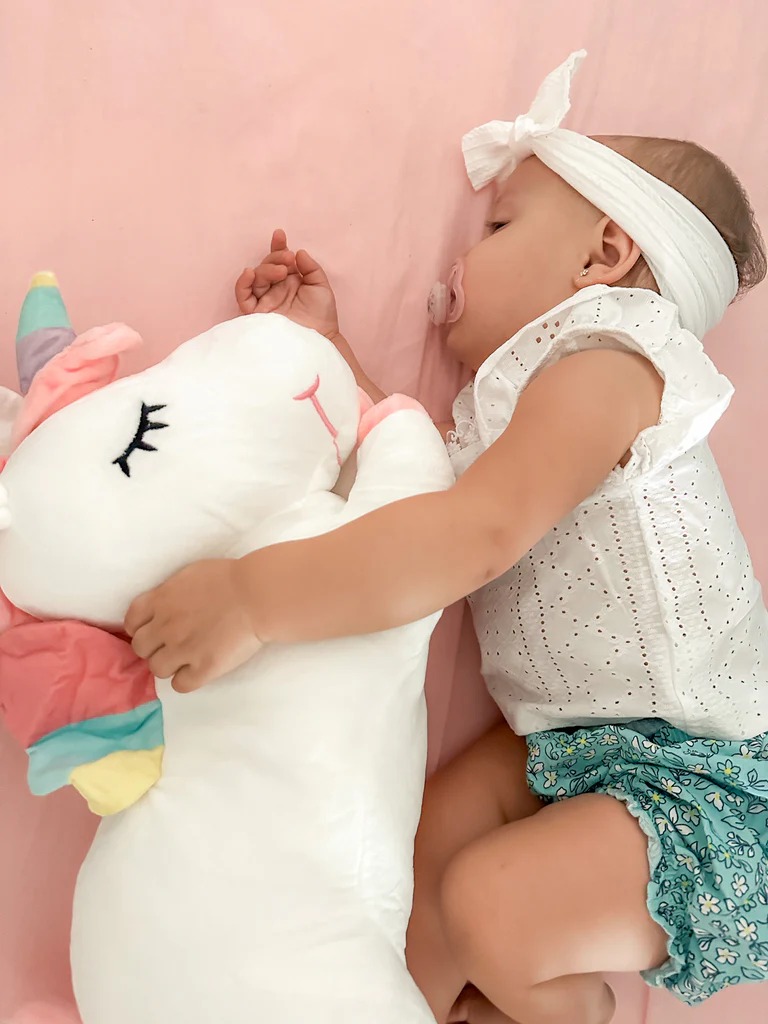 A baby girl wearing a white headband and pacifier sleeps peacefully on a pink sheet while hugging a large white unicorn plush toy with a colorful mane.