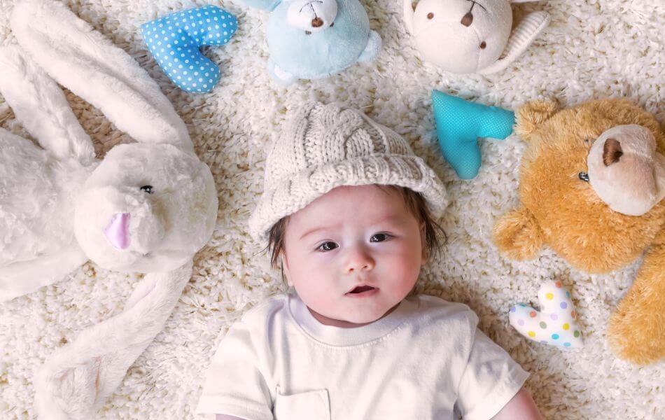 A baby lying on a soft carpet surrounded by plush toys, including a bunny, teddy bears, and heart-shaped cushions, wearing a knitted hat and a white onesie.