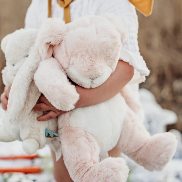 A child holding two large, fluffy stuffed bunnies—one pink and one white—while standing outdoors in soft lighting.