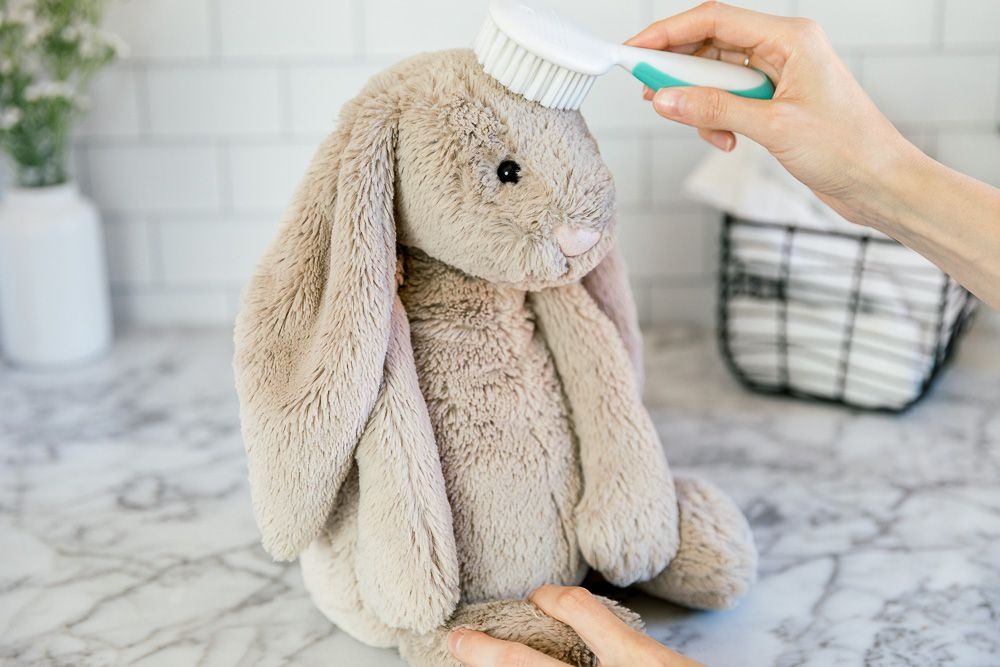 A person gently brushing a soft beige plush bunny toy with a white and green brush on a marble counter in a clean, tiled setting.