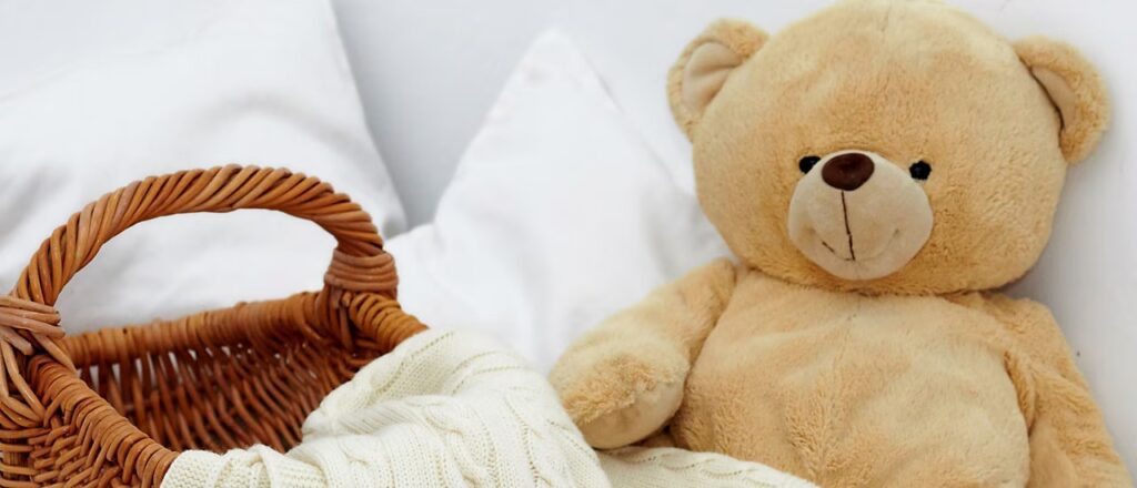 A soft beige teddy bear sitting next to a wicker basket filled with a cozy white knitted blanket, with white pillows in the background.