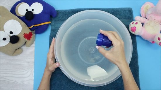 A person preparing to clean plush toys by pouring detergent into a large bowl of water, with three stuffed animals placed on a towel around the bowl.