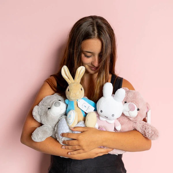 A young woman smiling and holding several plush toys, including a gray bear, tan bunny, white bunny, and pink bear, against a soft pink background.