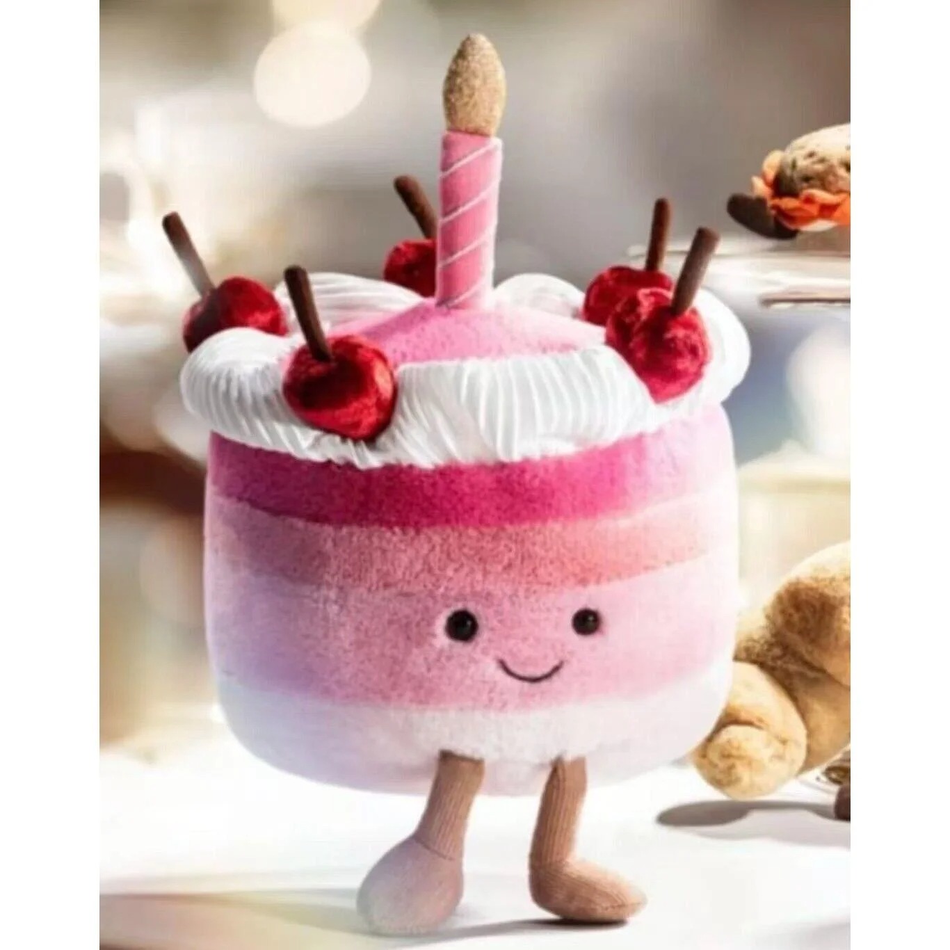 Plush pink birthday cake toy with smiling face, white icing top, red cherries, chocolate sticks, and a single candle on top, standing on two soft legs.