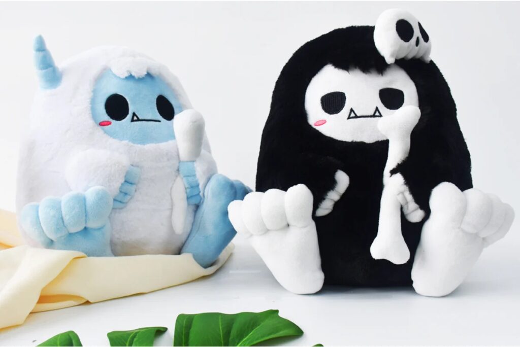 Two plush toys resembling cute monster characters, one in white and blue with a horn and the other in black and white with a skull hood, both holding soft bones and featuring fanged smiles and blush marks.