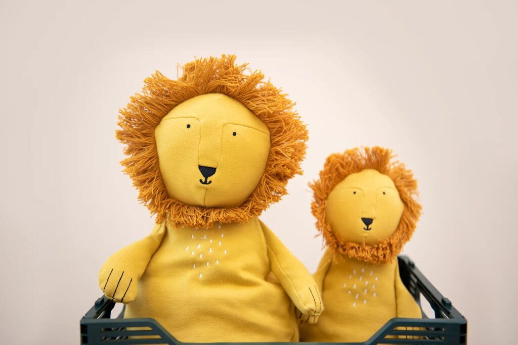 Two handmade plush lion toys with yellow bodies and orange yarn manes, sitting in a green crate against a light beige background.