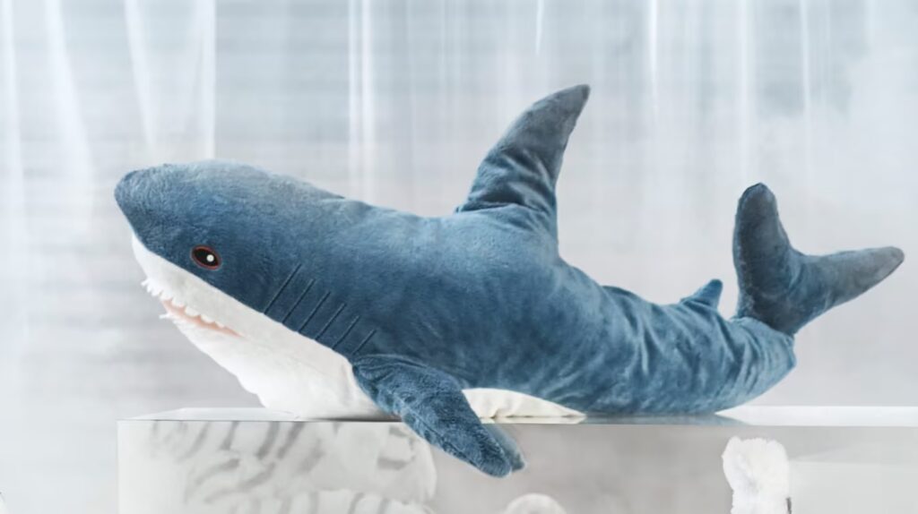 A large, blue plush shark toy with white underside and visible teeth, displayed on a white platform in a softly lit environment.