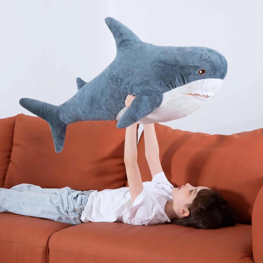A child lying on an orange couch holding up a large plush shark toy with both hands, playfully looking at it.