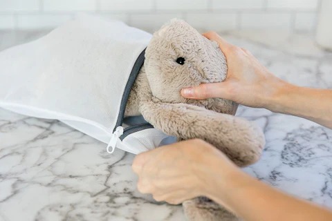 A person placing a plush bunny toy into a zippered mesh laundry bag on a marble surface, preparing it for safe machine washing.