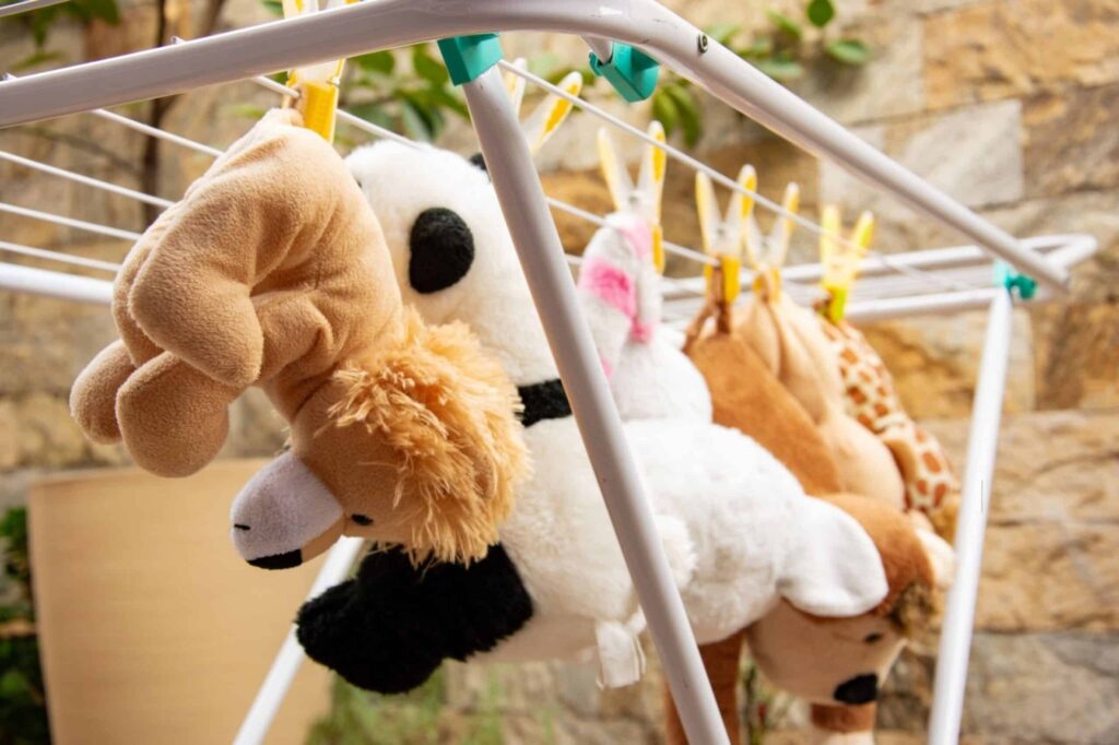 Several stuffed animals, including a panda, bear, and giraffe, hanging upside down with clothespins on a drying rack outdoors against a stone wall.