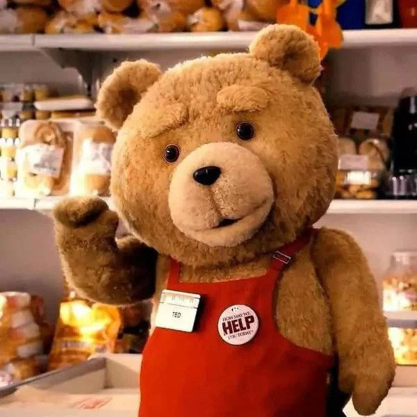 A plush teddy bear wearing a red apron and name tag that says "Ted," standing in a store aisle and waving, surrounded by packaged bread and snacks.