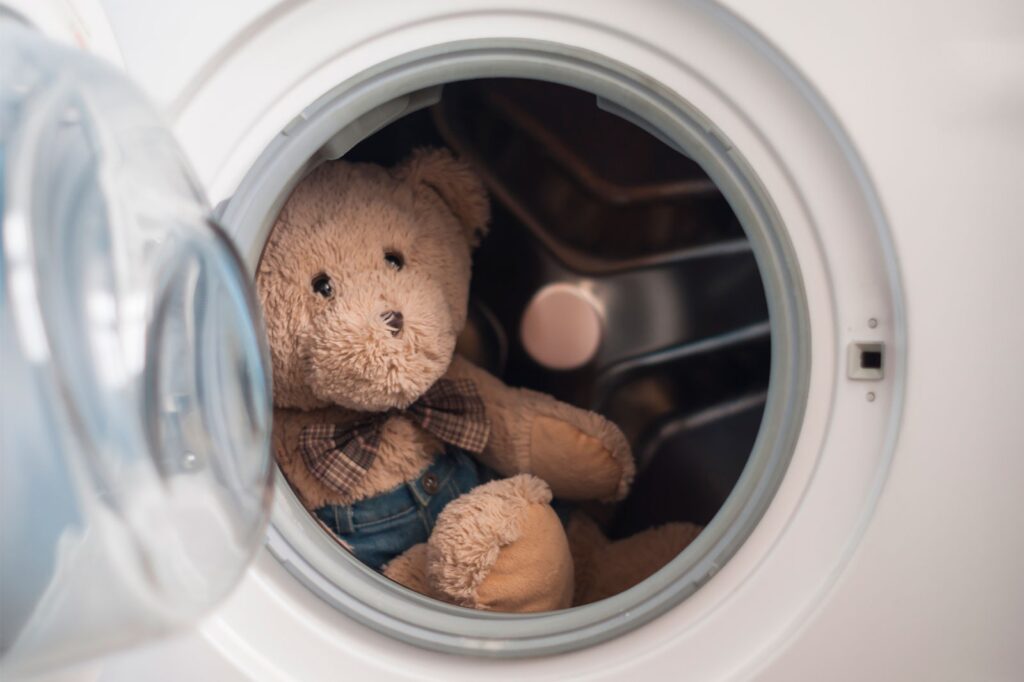 A brown teddy bear wearing a plaid bow tie and denim overalls sits inside the drum of a front-loading washing machine, ready to be cleaned.