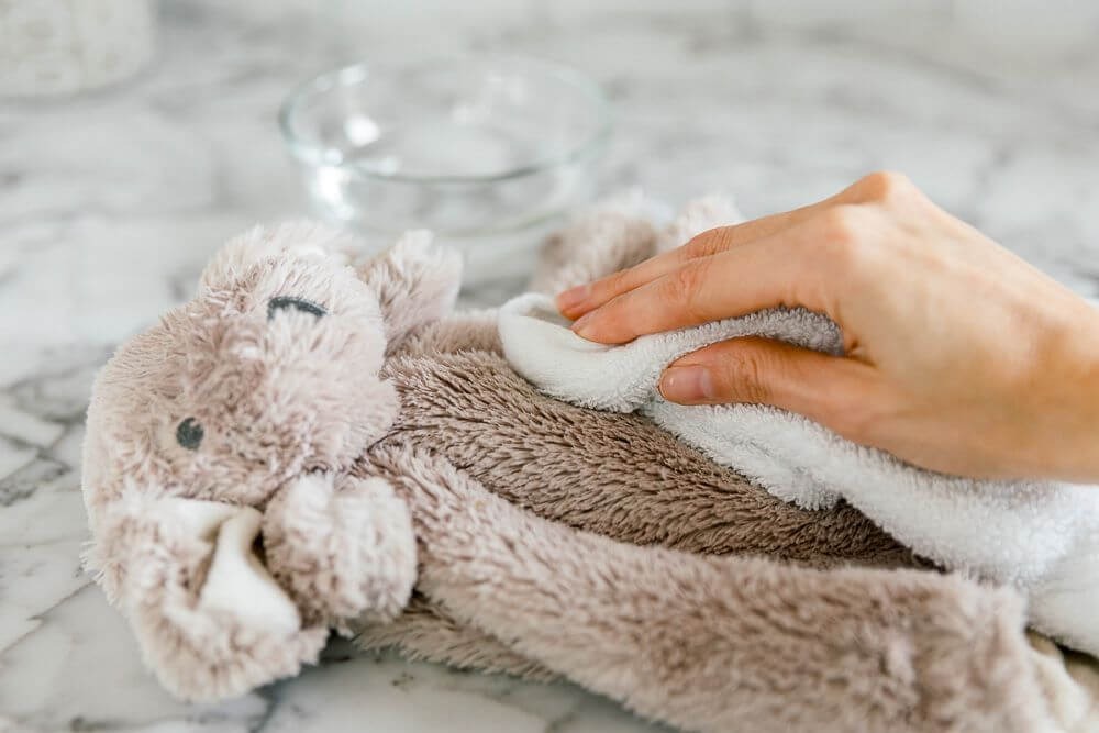 A person cleaning a soft beige plush bunny with a white cloth on a marble countertop, next to a glass bowl of water.