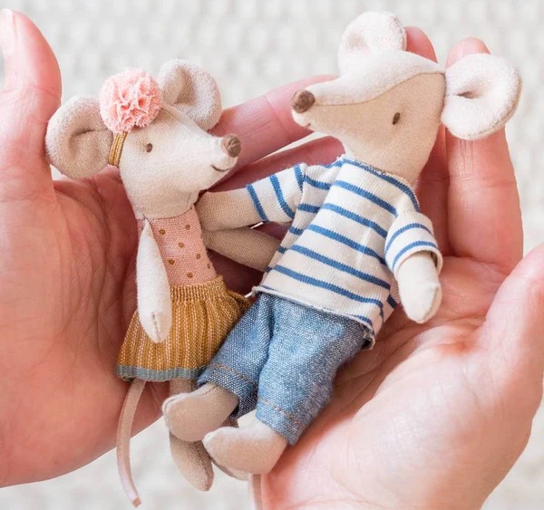Two small plush mice dolls wearing cute outfits held gently in hands, one in a dress with a flower headband and the other in a striped shirt and jeans.