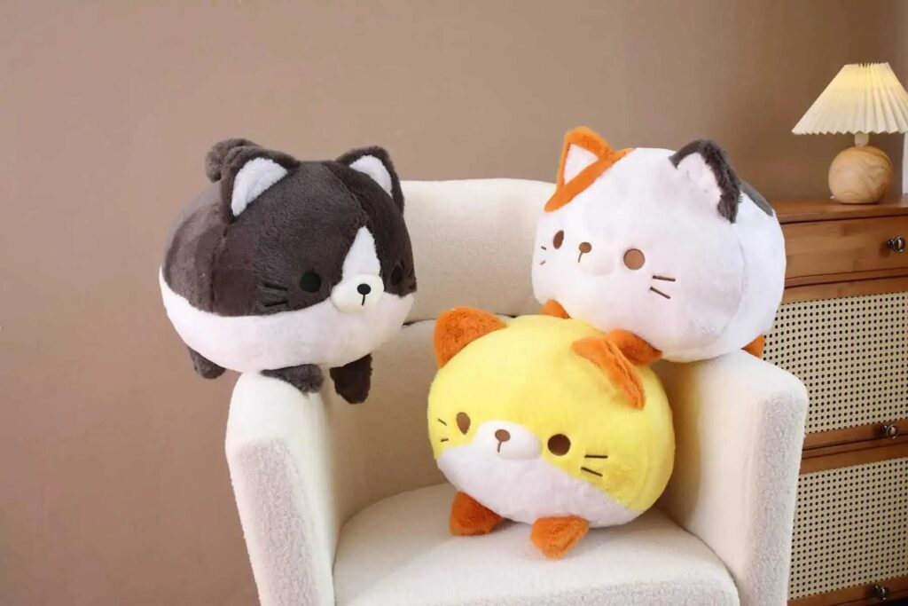 Three round cat plush toys in brown, yellow, and white sit on a white armchair in a cozy room.