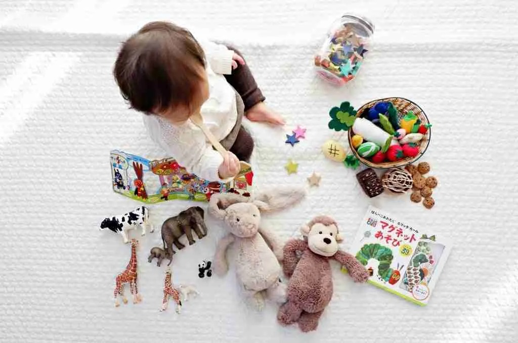 Baby sitting on a white blanket surrounded by plush toys, animal figurines, colorful felt stars, books, and a basket of play food.