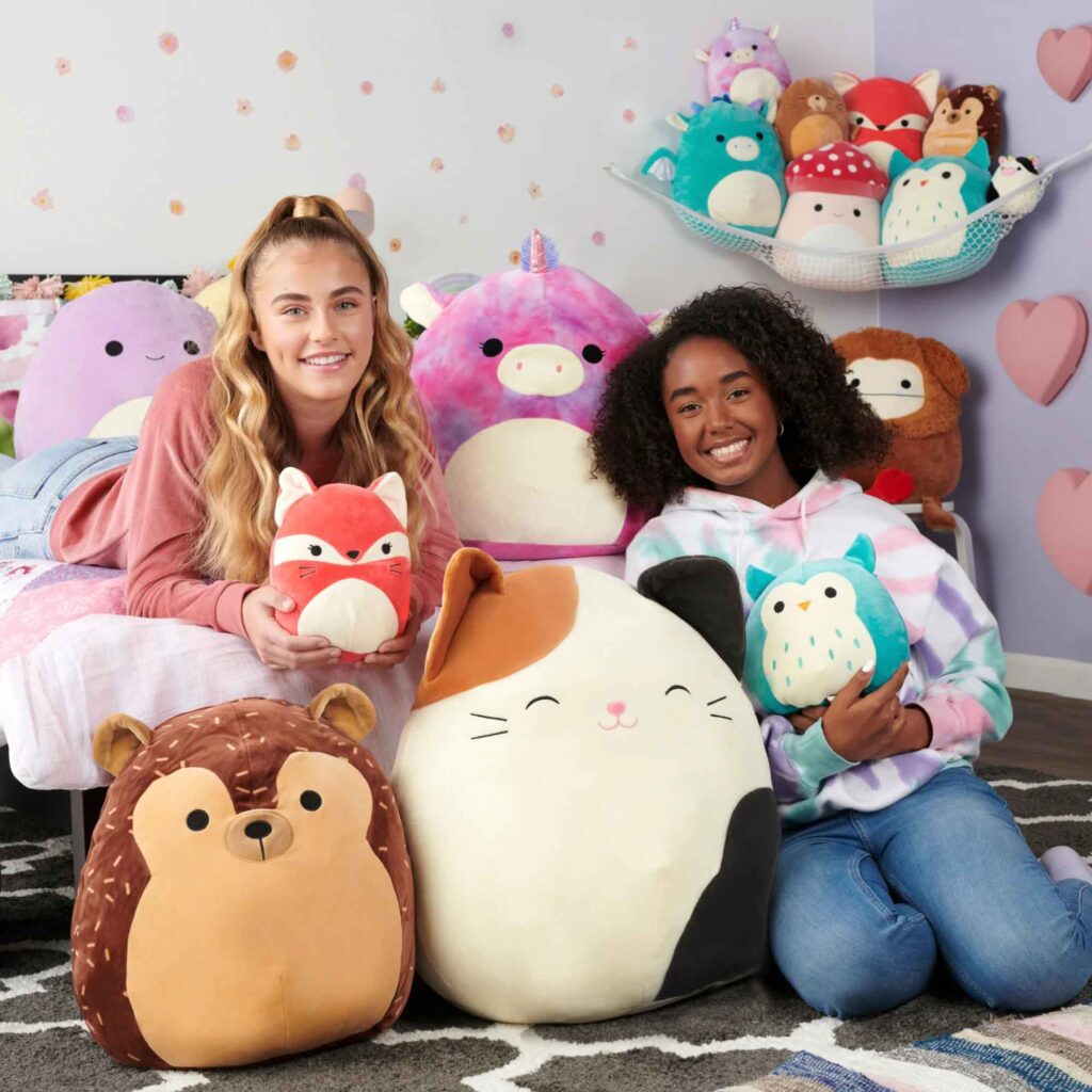 Two girls surrounded by colorful Squishmallow plush toys including a cat, fox, owl, unicorn, and bear in a cozy bedroom