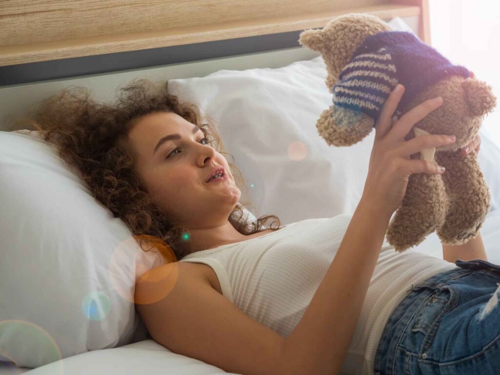 Smiling teenage girl lying on bed holding a plush teddy bear in striped sweater with morning sunlight
