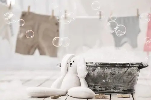 White plush bunny sitting next to a metal wash tub with soap bubbles and laundry hanging in the background