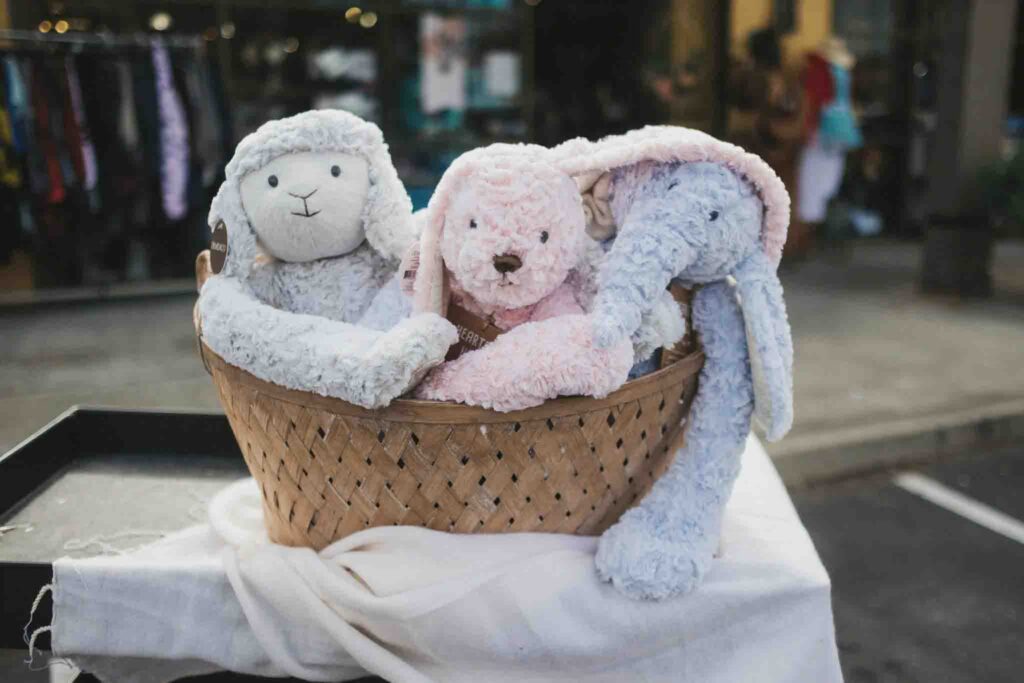 A wicker basket filled with soft plush toys including a gray lamb, pink puppy, and light blue elephant, displayed on a white cloth outdoors.