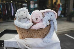 A wicker basket filled with soft plush toys including a gray lamb, pink puppy, and light blue elephant, displayed on a white cloth outdoors.