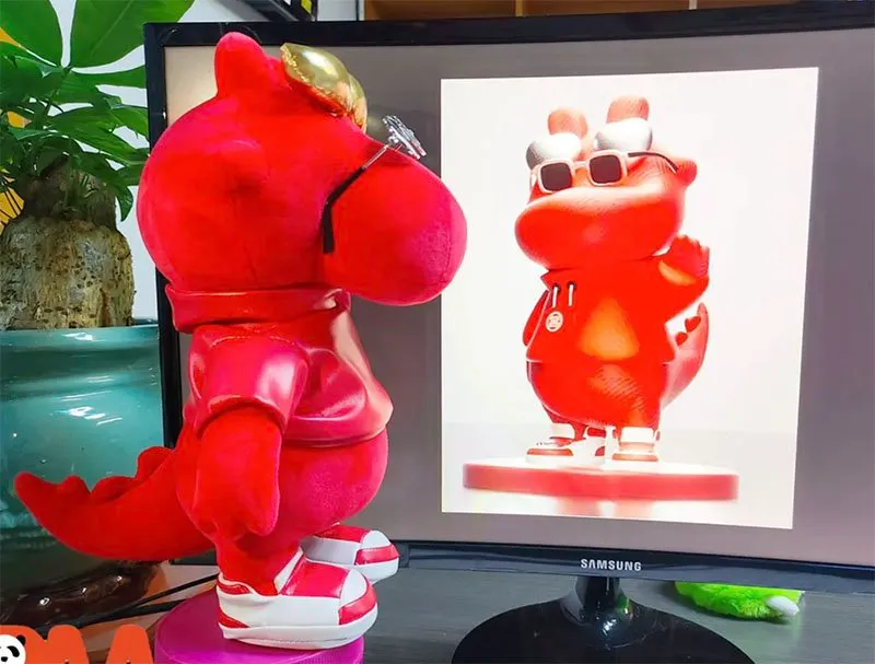 Red dinosaur plush toy wearing glasses and sneakers next to a screen showing its digital design.