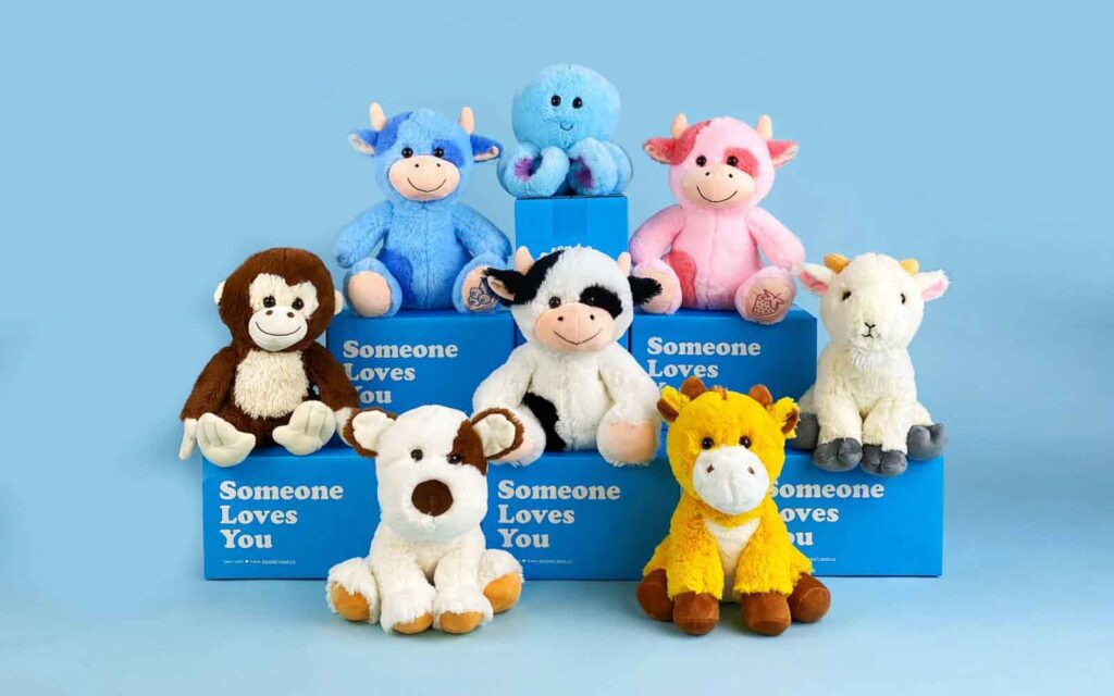 Group of colorful plush animals including cows, monkey, giraffe, octopus, goat, and dog on blue boxes with "Someone Loves You" text