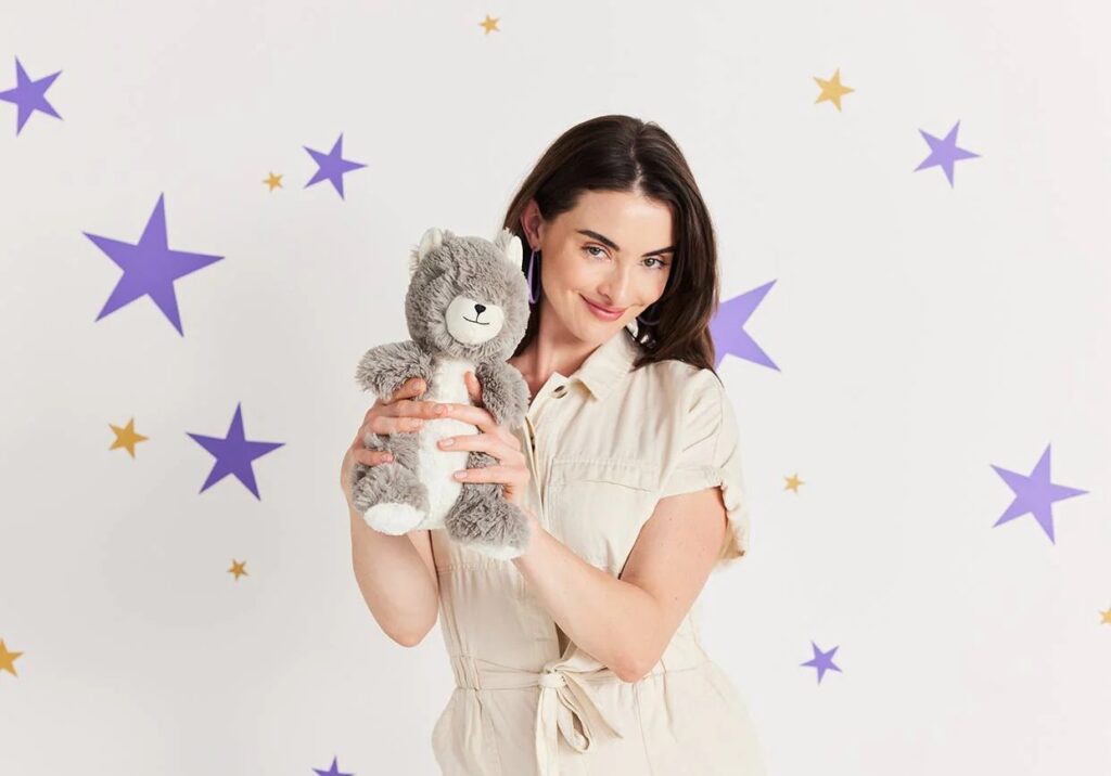 Uma mulher sorridente, com um fato bege claro, segurando um ursinho de peluche cinzento, encostada a uma parede decorada com decalques de estrelas roxas e douradas.