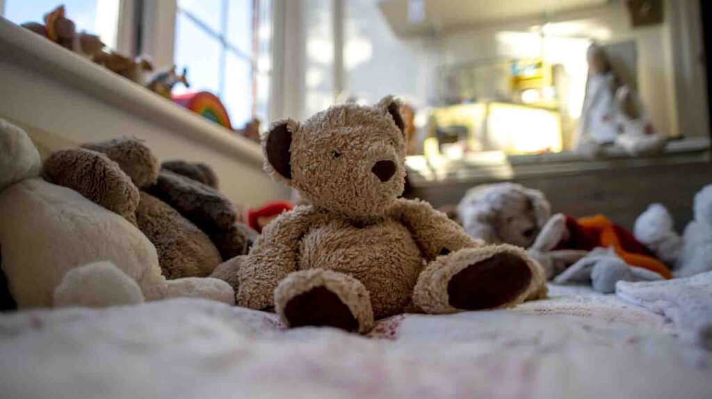 Un orsacchiotto marrone seduto in posizione verticale su un letto, circondato da altri peluche in una stanza illuminata da una luce soffusa.