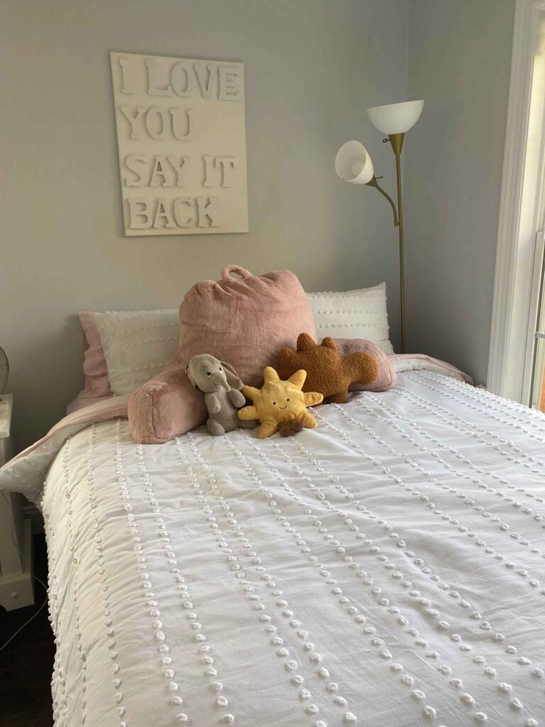 Plush toys including sun, elephant, and dinosaur on a white bed with textured bedding and a wall sign reading “I LOVE YOU SAY IT BACK”