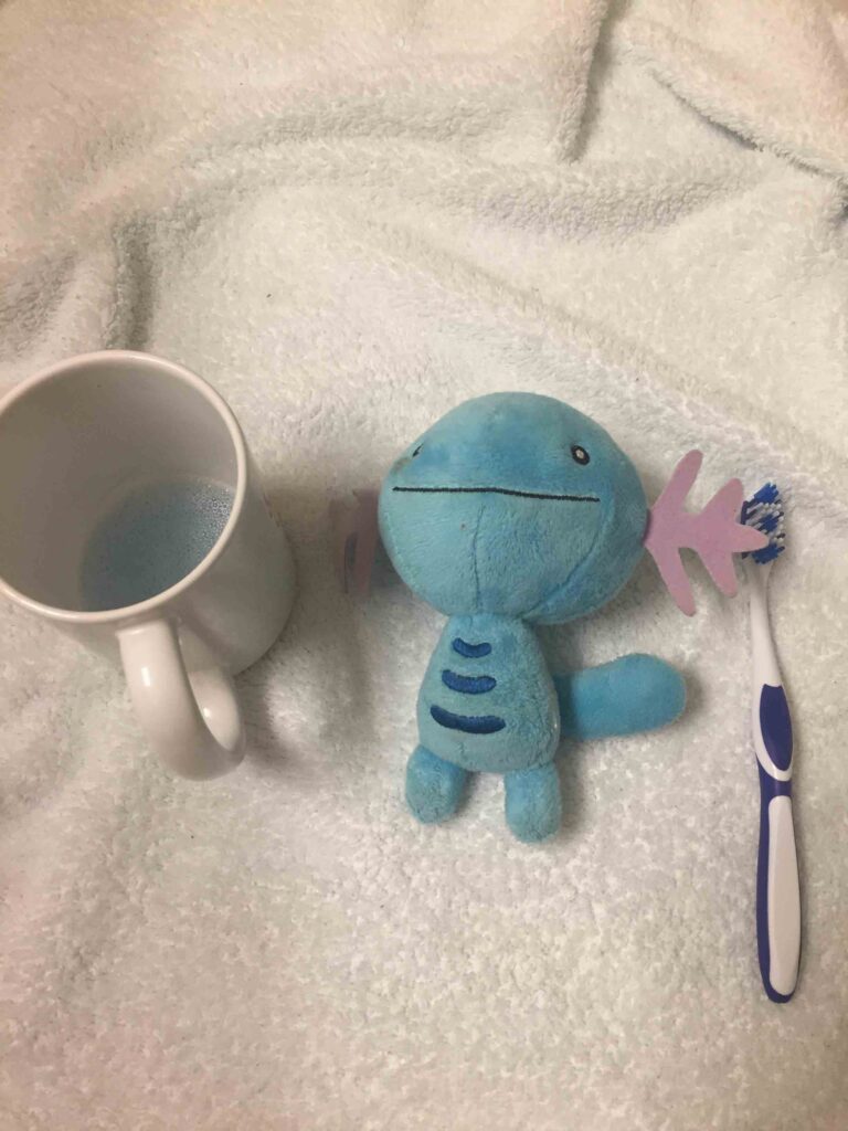 Blue plush toy placed on a towel beside a toothbrush and a white cup with mouthwash inside