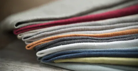Pilha de tecido de linho e algodão dobrado em cores variadas numa superfície de madeira.