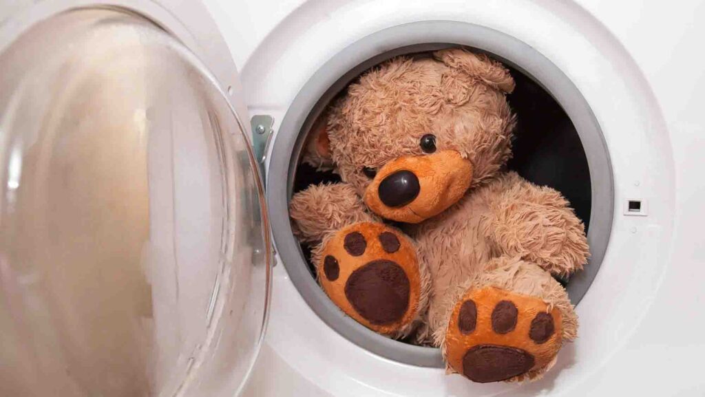 A fluffy brown teddy bear with orange paws inside a front-loading washing machine, ready for cleaning