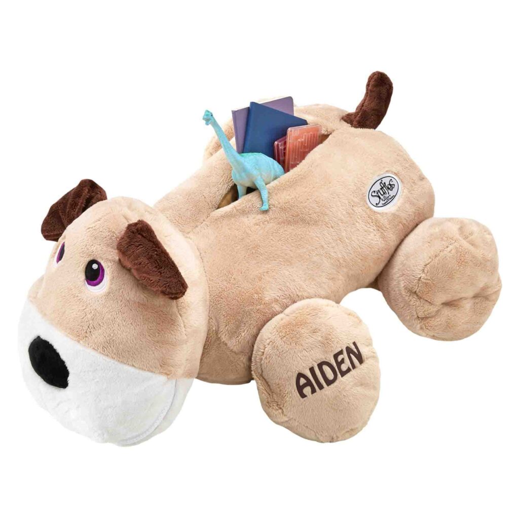 A tan plush dog with floppy ears labeled “AIDEN,” designed as a toy organizer with a storage pocket on its back holding small items like a dinosaur toy and books.