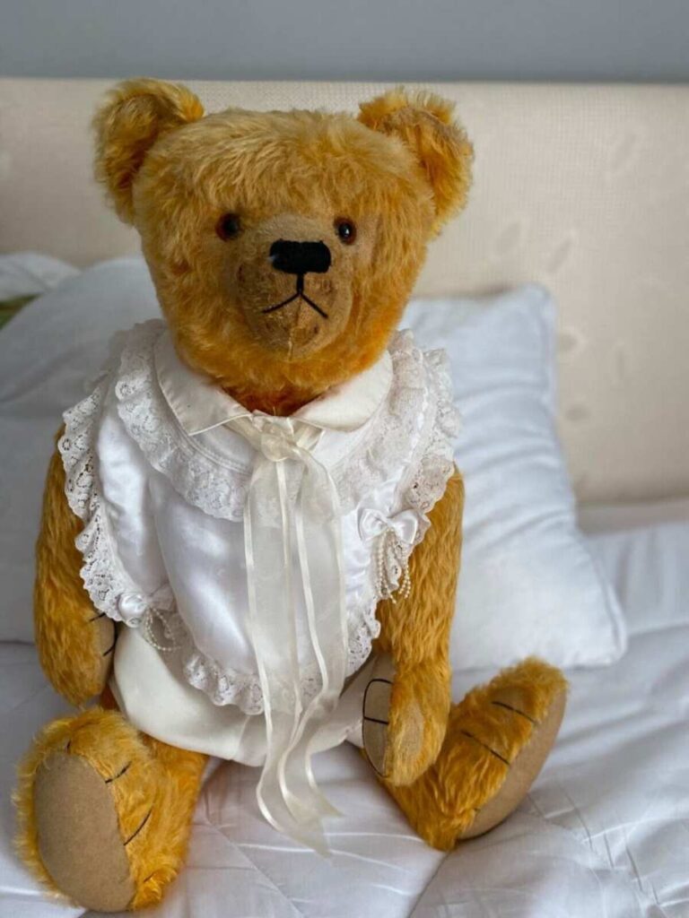 Ours en peluche de style ancien, vêtu d'une robe en dentelle blanche, assis sur un lit avec des oreillers.