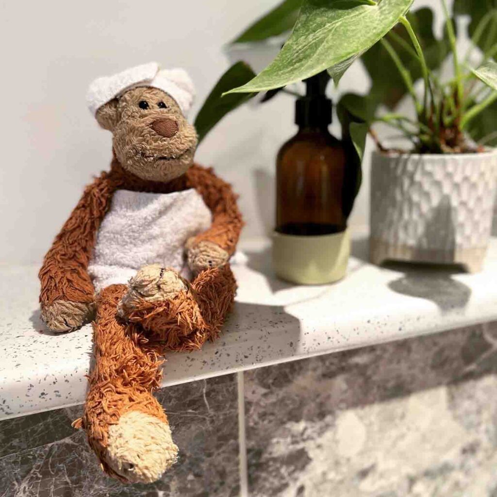 Una scimmia di peluche molto amata che indossa un asciugamano bianco e una fascia per la testa, seduta sul bancone del bagno accanto a una pianta e a un dispenser di sapone.
