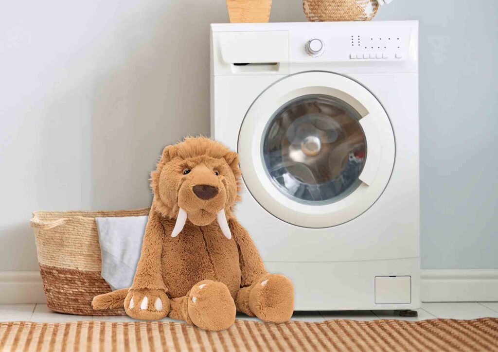 Cute brown plush mammoth with tusks sitting beside a white washing machine in a cozy laundry room