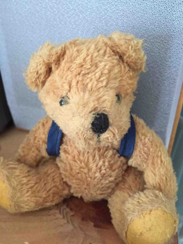 Vieil ours en peluche avec des bretelles bleues, assis sur une surface en bois.