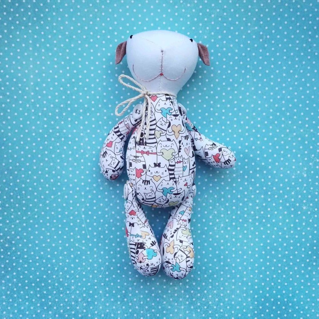 Um peluche de cão em tecido feito à mão, decorado com estampas de gatos de desenhos animados, sobre um fundo de bolinhas azuis com um laço de cordel à volta do pescoço.