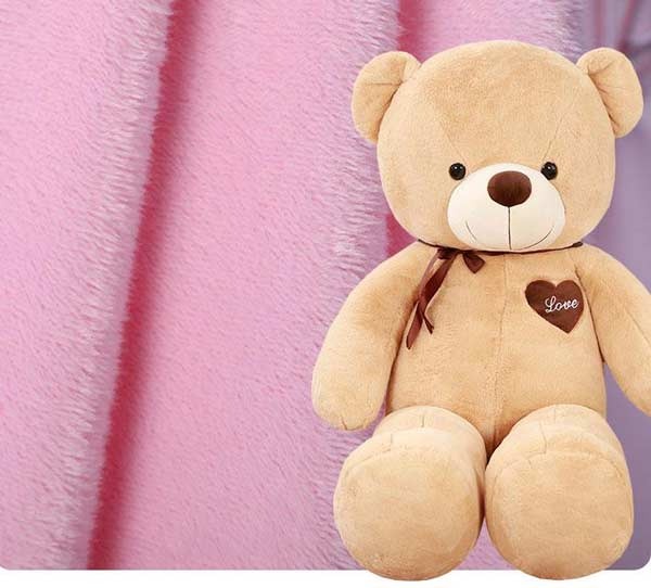 Um ursinho de peluche castanho com um coração com a inscrição "Love" sentado em frente a um fundo de tecido de lã cor-de-rosa suave.