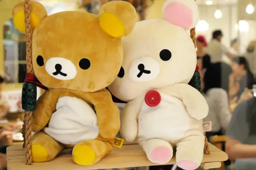 Brown and white Rilakkuma plush bears sitting on wooden swing indoors.
