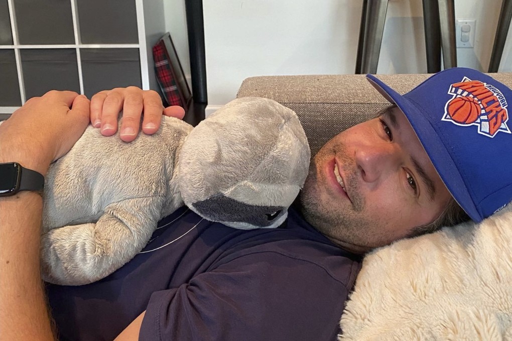 Smiling man wearing a Knicks cap lying on a couch and hugging a gray plush dinosaur toy
