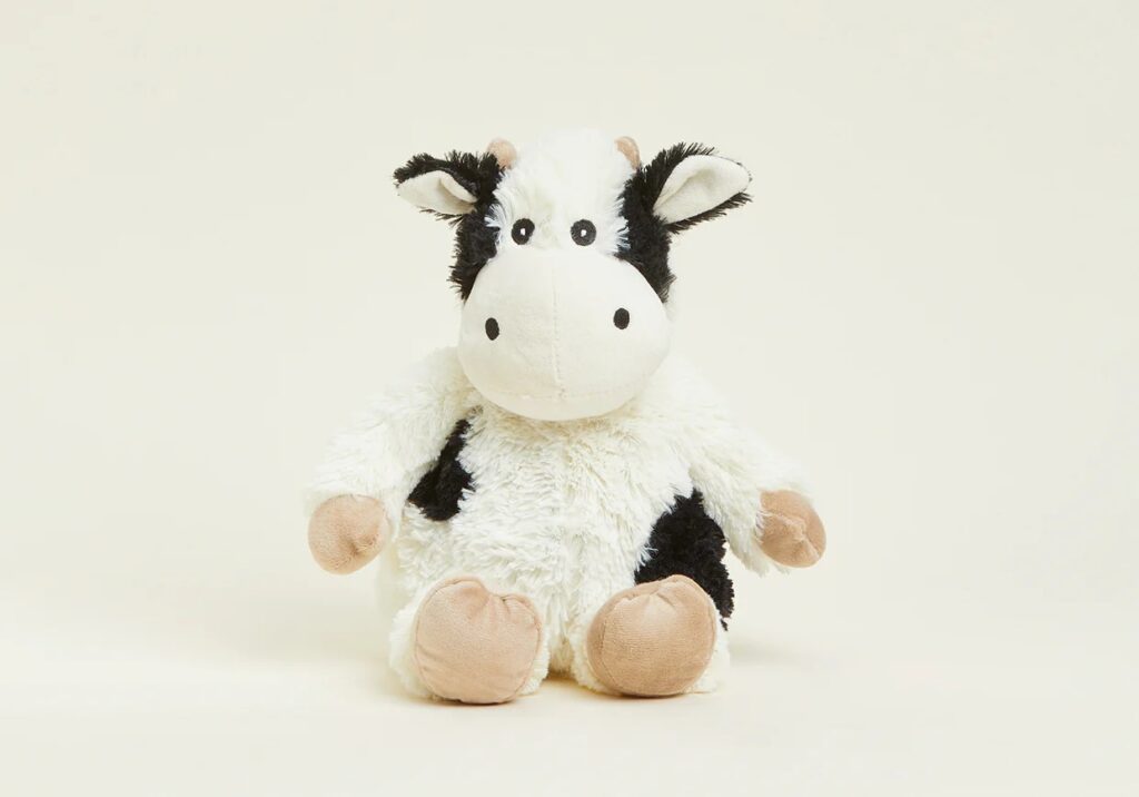 Uma vaca de peluche preta e branca macia com cascos e orelhas bege, sentada na vertical sobre um fundo branco liso.