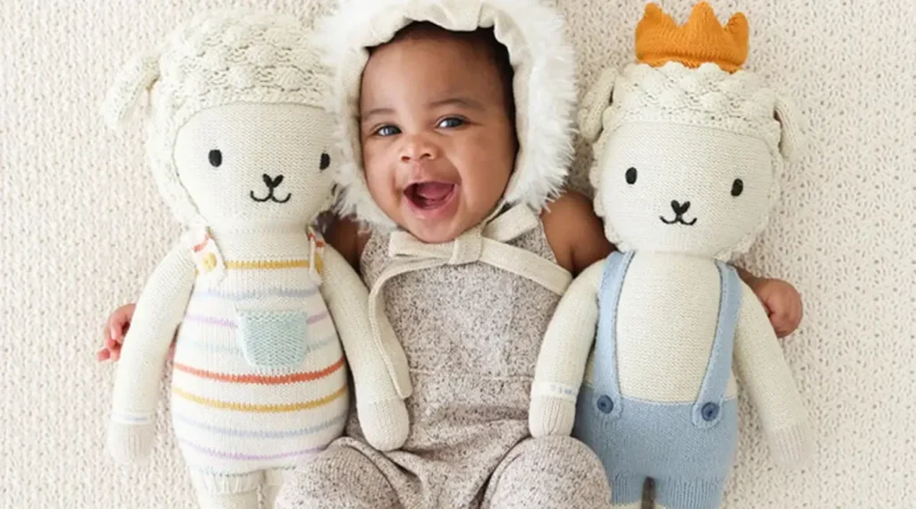 Um bebé feliz, vestido com um aconchegante fato com capuz, encontra-se entre dois peluches de animais de malha, um com um fato às riscas e outro com uma coroa e um macacão.
