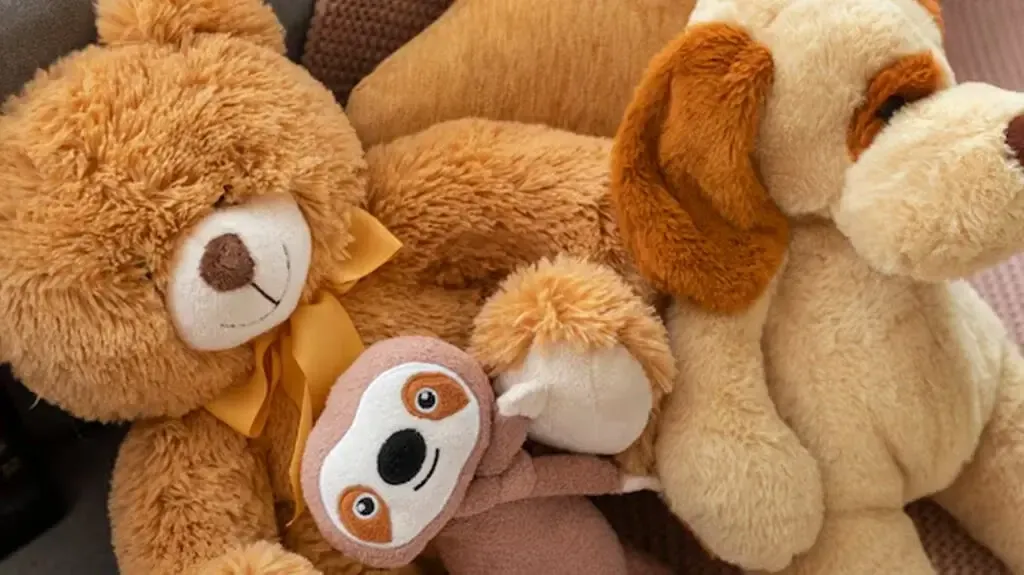 A teddy bear, a dog plush, and a smiling sloth toy snuggled together on a couch with warm-toned fabric.