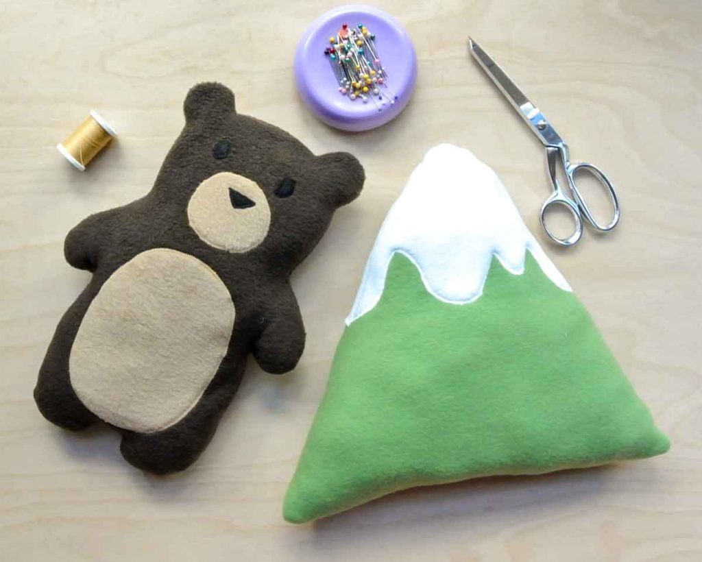 A handmade plush bear and mountain pillow toy placed on a wooden table with sewing supplies including scissors, thread, and pins.