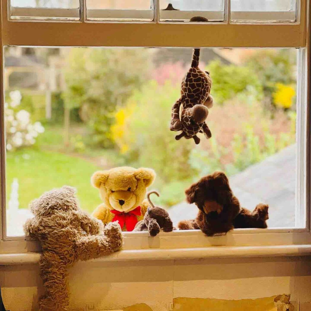Diversi animali di peluche, tra cui orsacchiotti, una giraffa e un topo, sono disposti in modo giocoso sul davanzale di una finestra con un giardino verde sullo sfondo.