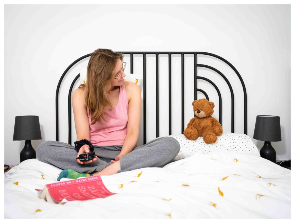 Woman sitting on bed with remote control looking at a plush teddy bear placed on a pillow beside her